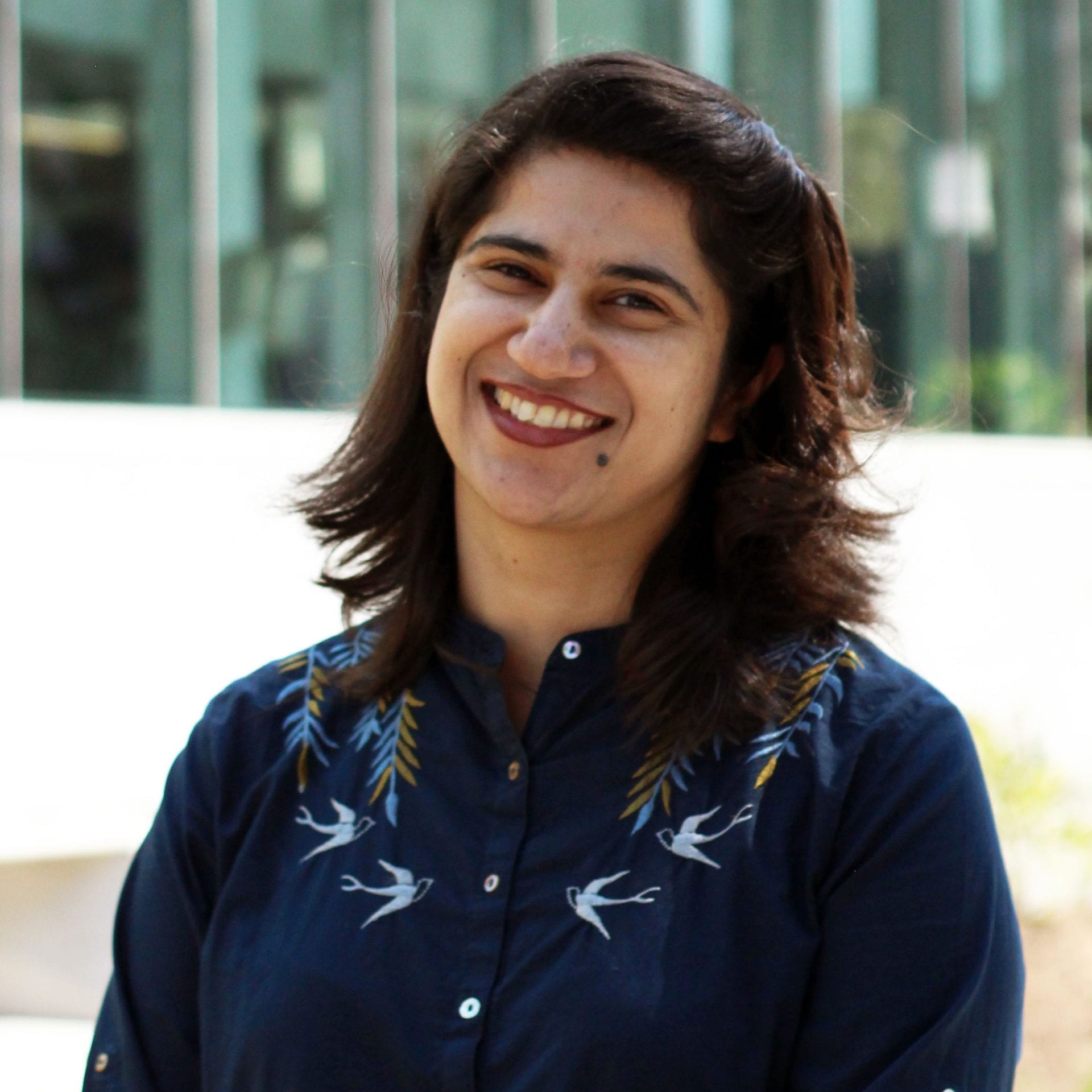 mehr-mahzoor A person smiling for the camera, wearing a blue shirt standing in front of a white building with windows.