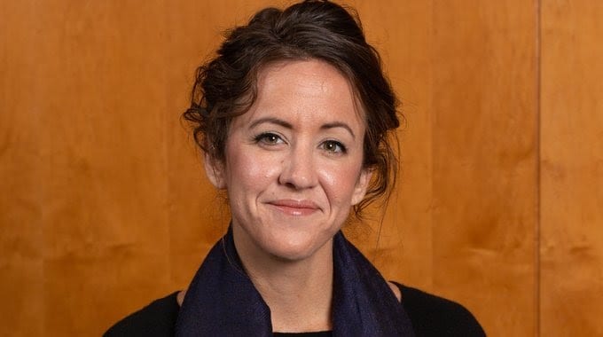 Kelly Orians smiling wearing a black shirt in front of a wooden wall.