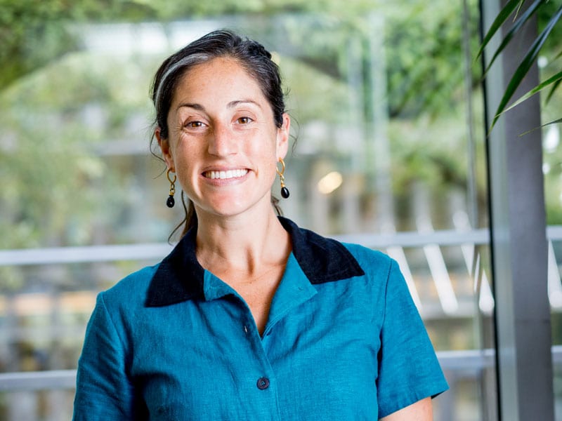 Woman Smiling Wearing A Blue Shirt With A Black Collar In Front Of Trees.